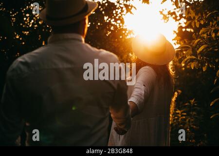 Una coppia caucasica vestita di bianco e con cappelli si posano in un giardino pieno di alberi che camminano mano in mano Foto Stock
