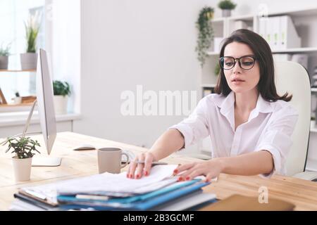 Attraente giovane donna d'affari seduto alla scrivania in ufficio e trovare documenti in stack Foto Stock