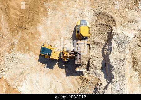 Pala caricatrice della benna caricando la ghiaia su un rimorchio di autocarro articolato in un cantiere di grandi dimensioni, vista dall'alto in basso. Foto Stock