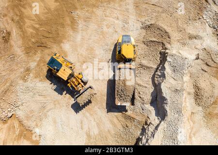 Pala caricatrice della benna caricando la ghiaia su un rimorchio di autocarro articolato in un cantiere di grandi dimensioni, vista dall'alto in basso. Foto Stock