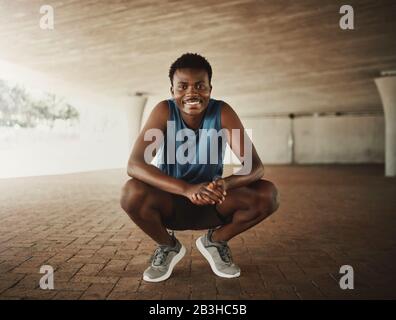 Ritratto di un felice afroamericano fitness giovane corridore maschio accovacciato sul marciapiede sorridente e guardando la macchina fotografica Foto Stock