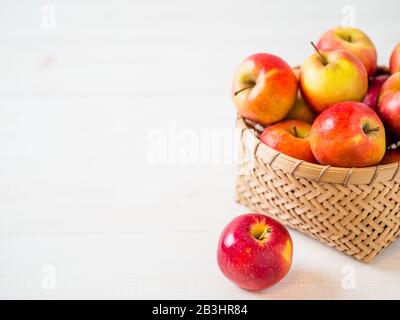 Concetto di raccolta delle mele. Cesto di bambù intrecciato con mele rosse su sfondo bianco di legno con spazio di copia per il testo o il design. Foto Stock