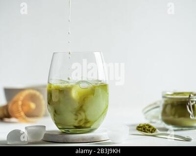 Tè Matcha latte ghiacciato con gocce di latte versato in bicchiere. Latte Matcha e ingredienti su sfondo di marmo bianco. Spazio di copia per txt o design Foto Stock