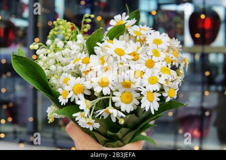 Femmina mano che tiene bouquet di margherite e gigli della valle contro una città luci sfondo Foto Stock