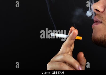 Un giovane tiene una sigaretta nelle mani e esala il fumo su uno sfondo scuro. Danno da fumo nicotina e tabacco usare da vicino. Foto Stock