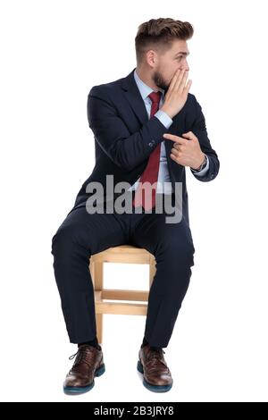 felice giovane uomo d'affari che sussurra e punta le dita, guardando di lato e seduto isolato su sfondo bianco in studio Foto Stock