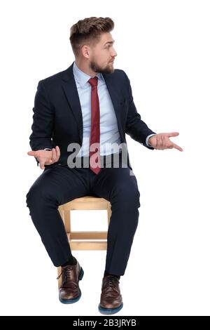 giovane uomo d'affari confuso che guarda da parte, tenendo le mani e gesticolando, parlando, seduto isolato su sfondo bianco in studio Foto Stock