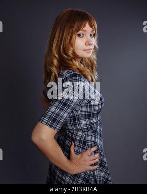 Una ragazza con ricci dorati sembra mezzo giro. Indossa un abito blu a scacchi. La mano si trova sull'anca. Studio. Foto Stock