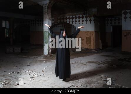 donna bruna gotica alzando le mani in piedi in grande sala vuota di casa oscura abbandonata Foto Stock