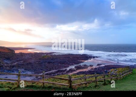 Il sole si rompe attraverso le nuvole tempesta, una recinzione di legno in primo piano conduce l'occhio verso la spiaggia dove le onde si rompono sulla spiaggia e rocce. Foto Stock