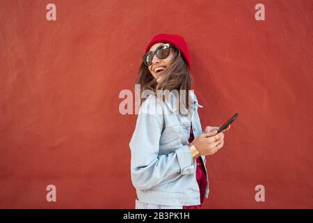 Ritratto di una giovane e allegra donna elegantemente vestita di cappello e giacca in piedi con telefono cellulare sulla parete rossa di fondo all'aperto Foto Stock