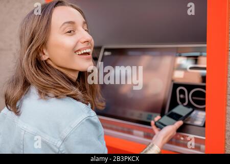 Giovane donna che ritira contanti da un bancomat utilizzando un telefono cellulare sulla strada all'aperto Foto Stock