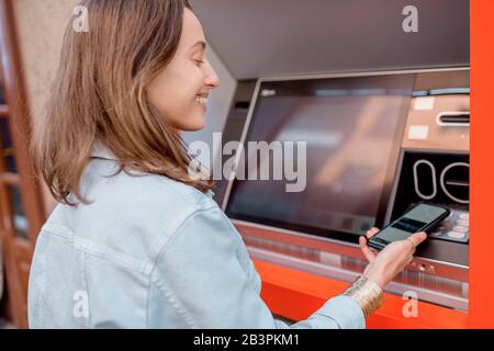 Giovane donna che ritira contanti da un bancomat utilizzando un telefono cellulare sulla strada all'aperto Foto Stock