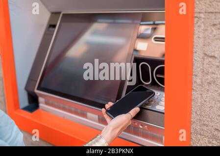 Giovane donna che ritira contanti da un bancomat utilizzando un telefono cellulare sulla strada all'aperto, primo piano sul telefono Foto Stock