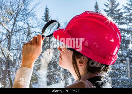 fuoco selettivo su donna ispettore che indossa un cappello duro rosa, lei usando una lente d'ingrandimento sulla finestra. con alberi innevati all'aperto sullo sfondo. Foto Stock