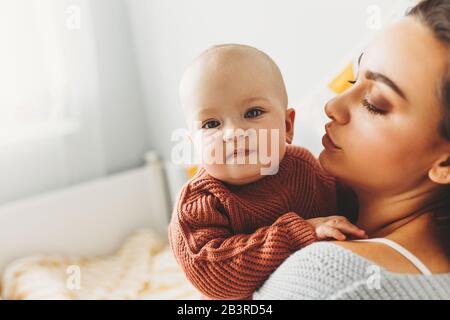 Una bambina sorride e gioca sul letto con la mamma, vestita con maglioni in maglia tonalità pastello alla moda, in una stanza, casa accogliente, design moderno. Famiglia felice a casa, concetto di maternità Foto Stock