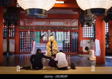 Il sacerdote Shinto apparentemente benedice il giovane ragazzo seduto tra due adulti in vista pubblica, nel Santuario di Marodo all'interno del complesso Santuario di Itsukushima, Miyajima Foto Stock