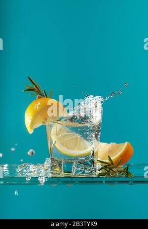 Cocktail gin-tonic con limone e rosmarino. Bevanda gassata con pezzi di ghiaccio su un tavolo di vetro. Foto Stock