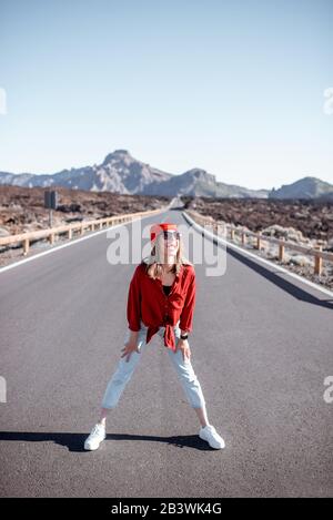 Ritratto di stile di vita di una giovane donna elegantemente vestita di rosso a piedi sulla bella strada in mezzo alla valle vulcanica in una giornata di sole. Stile di vita spensierato e concetto di viaggio Foto Stock