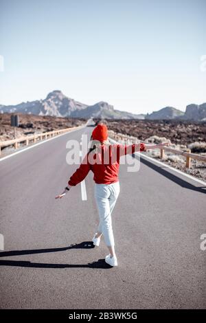 Ritratto di stile di vita di una giovane donna elegantemente vestita di rosso a piedi sulla bella strada in mezzo alla valle vulcanica in una giornata di sole. Stile di vita spensierato e concetto di viaggio Foto Stock