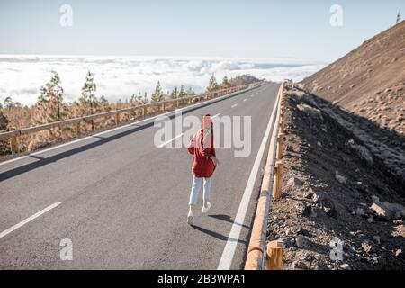 Ritratto di stile di vita di una giovane donna elegantemente vestita in rosso correndo sulla bella strada sopra le nuvole in una giornata di sole. Stile di vita spensierato e concetto di viaggio Foto Stock