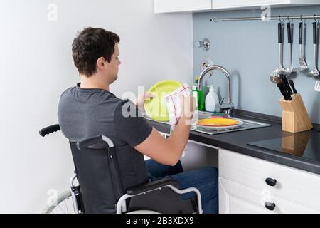 Uomo disabile seduto su una sedia a rotelle per il lavaggio e la pulizia delle stoviglie in cucina Foto Stock