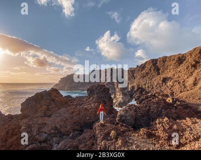 Donna che gode di una splendida vista sulla costa rocciosa dell'oceano e un faro sullo sfondo. Immagine realizzata sul telefono cellulare Foto Stock