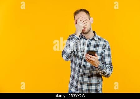 Ritratto di studente stanco maschio isolato su giallo utilizzando il suo smartphone. Facepalm. Foto Stock