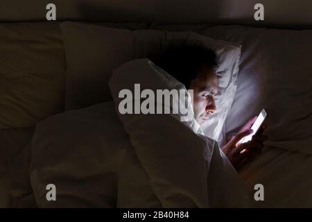 Dipendenza mobile, nomofobia, insonnia, concetto di disturbo del sonno. L'uomo annoiato che chiacchierava e naviga su internet con lo smartphone in tarda notte, sdraiato su BE Foto Stock