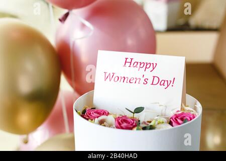 Testo Happy Womens Day su carta regalo in fiore scatola vicino rosa festiva e palloncini d'oro. Biglietto d'auguri per le donne l'8 marzo, Giornata Internazionale della Donna Foto Stock