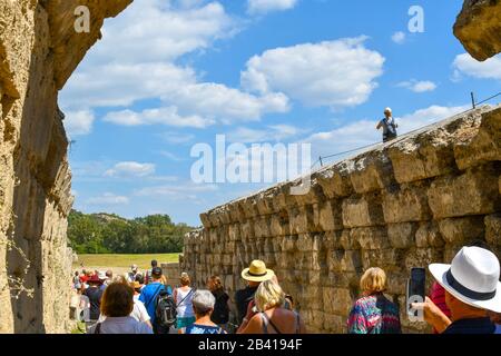 Un turista femminile si affaccia sull'antico stadio dell'arena nella città di Olimpia, in Grecia, mentre i turisti entrano sotto un'antica arcata Foto Stock