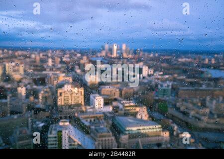 Gocce di pioggia su una finestra di vetro con sfondo sfocato del Fiume Tamigi e del quartiere finanziario di City of London Canary Wharf Foto Stock