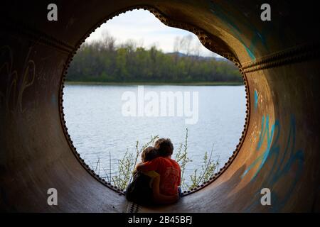 Due giovani fratelli si siedono insieme e abbracciano all'interno di un vuoto tubo di scarico arrugginito che si affaccia su un lago in un parco naturale a Windsor, Sonoma County, California. Foto Stock