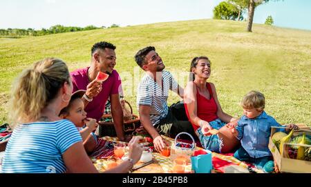 Multirazziale famiglie felici facendo picnic in parco naturale all'aperto - giovani genitori divertirsi con i bambini in estate - concetto di cibo e amicizia - Foto Stock