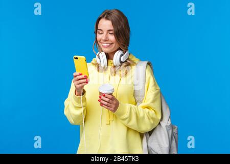 Ragazza che usa il telefono cellulare per contattare l'amico dopo l'università, scrivere come camminare lungo la strada con zaino, prendere caffè tazza e cuffie, sorridendo Foto Stock