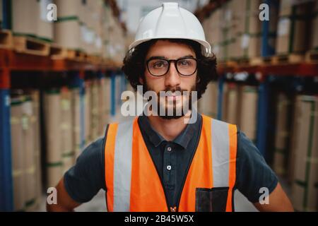 Ritratto di giovane lavoratore caucasico fiducioso in elmetto bianco e tuta protettiva con occhiali in piedi in negozio industriale Foto Stock