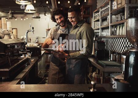 Sorridente bel cameriere che tiene un tablet con un collega mentre punta su di esso nella caffetteria che condivide la ricetta Foto Stock