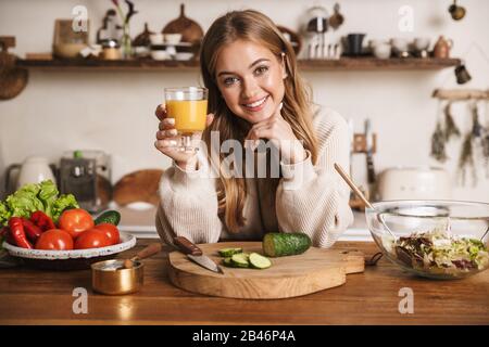 Immagine di donna sorridente cute che indossa abiti casual bere succo mentre si fa il pranzo in cucina accogliente Foto Stock