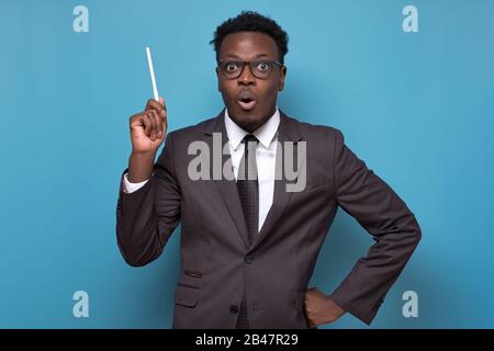 Stupito stupefied bell'uomo africano apre ampiamente la bocca, alza la penna in su come ottiene l'idea o ricorda di fare qualcosa non nel tempo. Studio girato su Blue wal Foto Stock