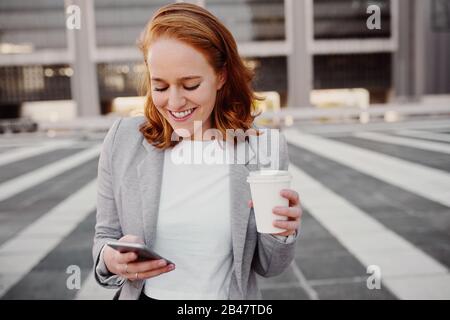 Ritratto di giovane donna di successo di affari che usa lo smartphone in piedi all'aperto che tiene takeaway tazza di caffè Foto Stock