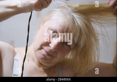 Donna anziana asciuga i capelli con un asciugacapelli a casa. Foto Stock
