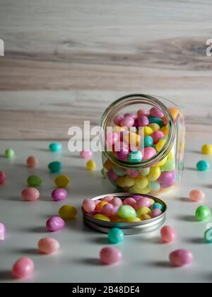 Fagioli di gelatina color pastello in un vaso di vetro che spargendo fuori su un contatore bianco con uno sfondo di legno. Caramelle e colori di Pasqua. Foto Stock