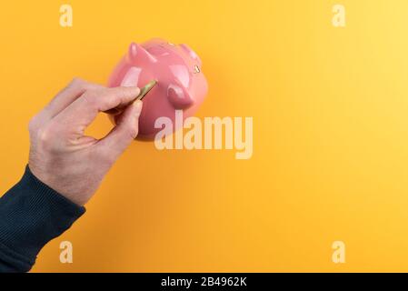 risparmio di denaro e il concetto di profitto, vista dall'alto della mano inserendo moneta dorata in banca rosa piggy contro sfondo arancione colorato Foto Stock