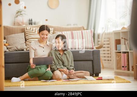 Due ragazze asiatiche seduti insieme sul pavimento contro il divano che guarda la carta di vacanza fatta a mano sorridente, scatto orizzontale, spazio copia Foto Stock