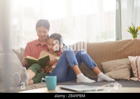 Orizzontale scatto di matura donna asiatica rilassante sul divano con la sua bella figlia lettura interessante libro il giorno del fine settimana Foto Stock