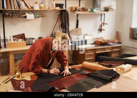 Colpo orizzontale di giovane craftswoman caucasico in piedi a tavola in officina foratura in cuoio nero parti di materiale per mettere rivetti Foto Stock