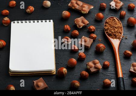 Cioccolato con nocciole, un taccuino con pagine bianche e un cucchiaio di legno con cacao su sfondo scuro, circondato da noci nel guscio e sbucciato. Foto Stock