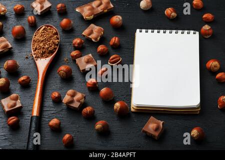 Cioccolato con nocciole, un taccuino con pagine bianche e un cucchiaio di legno con cacao su sfondo scuro, circondato da noci nel guscio e sbucciato. Foto Stock