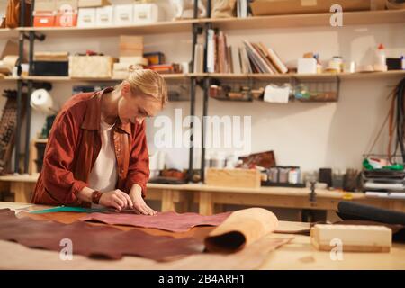 Colpo orizzontale di giovane donna caucasica che taglia fuori materiale di cuoio in officina moderna, spazio della copia Foto Stock