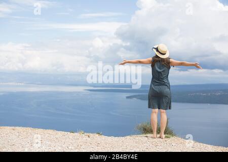 Donna in abito di lino e cappello è in piedi sulla costa della Croazia, diffonde le braccia e guarda al mare Foto Stock